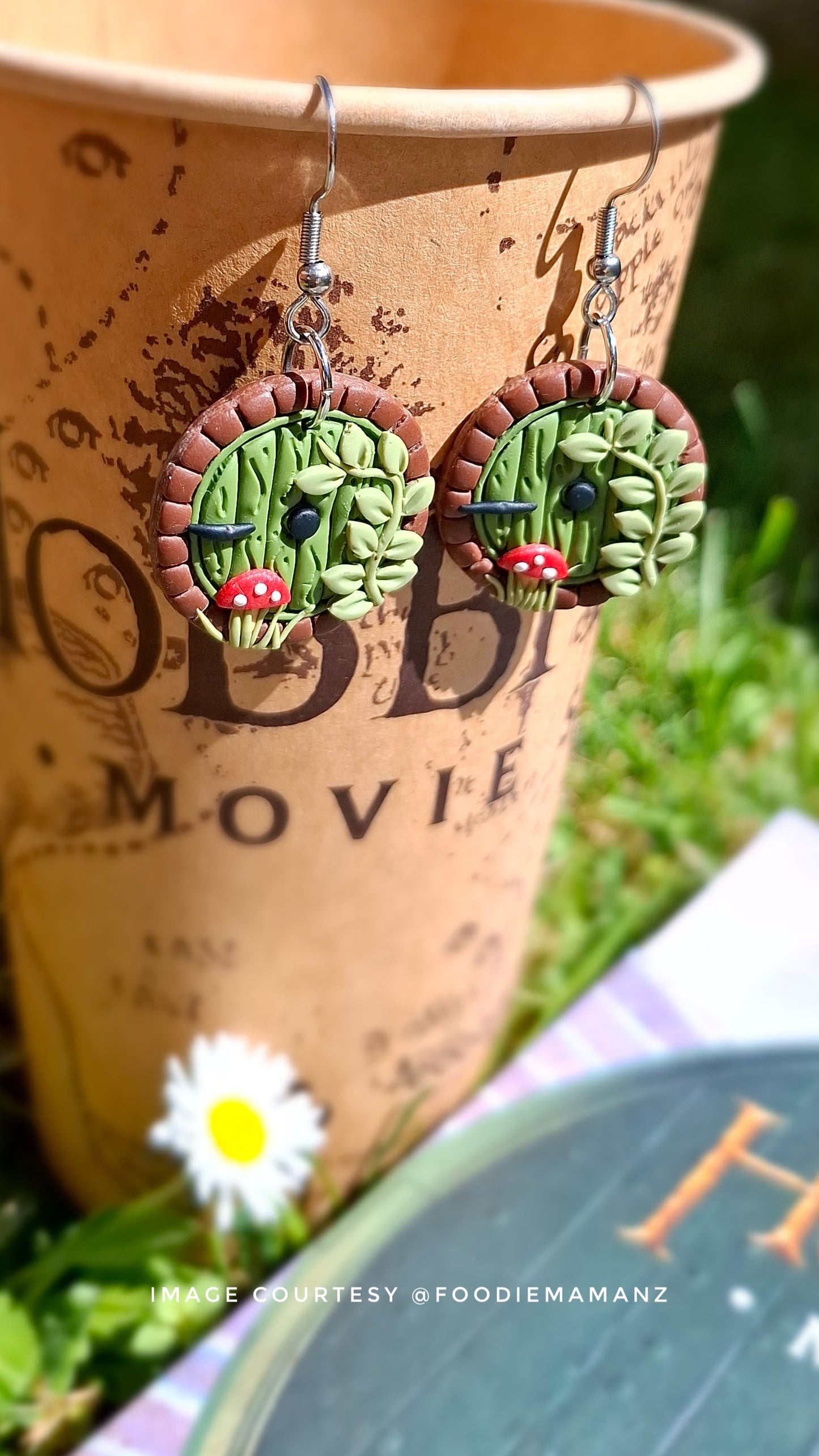 Close-up of Hobbit Door Polymer Clay Earrings by silverstar.nz, crafted with green doors and red mushrooms. They are displayed on a brown paper cup marked MOVIE, with a daisy on green grass beside an open book. Image credit to @foodiemamanz.
