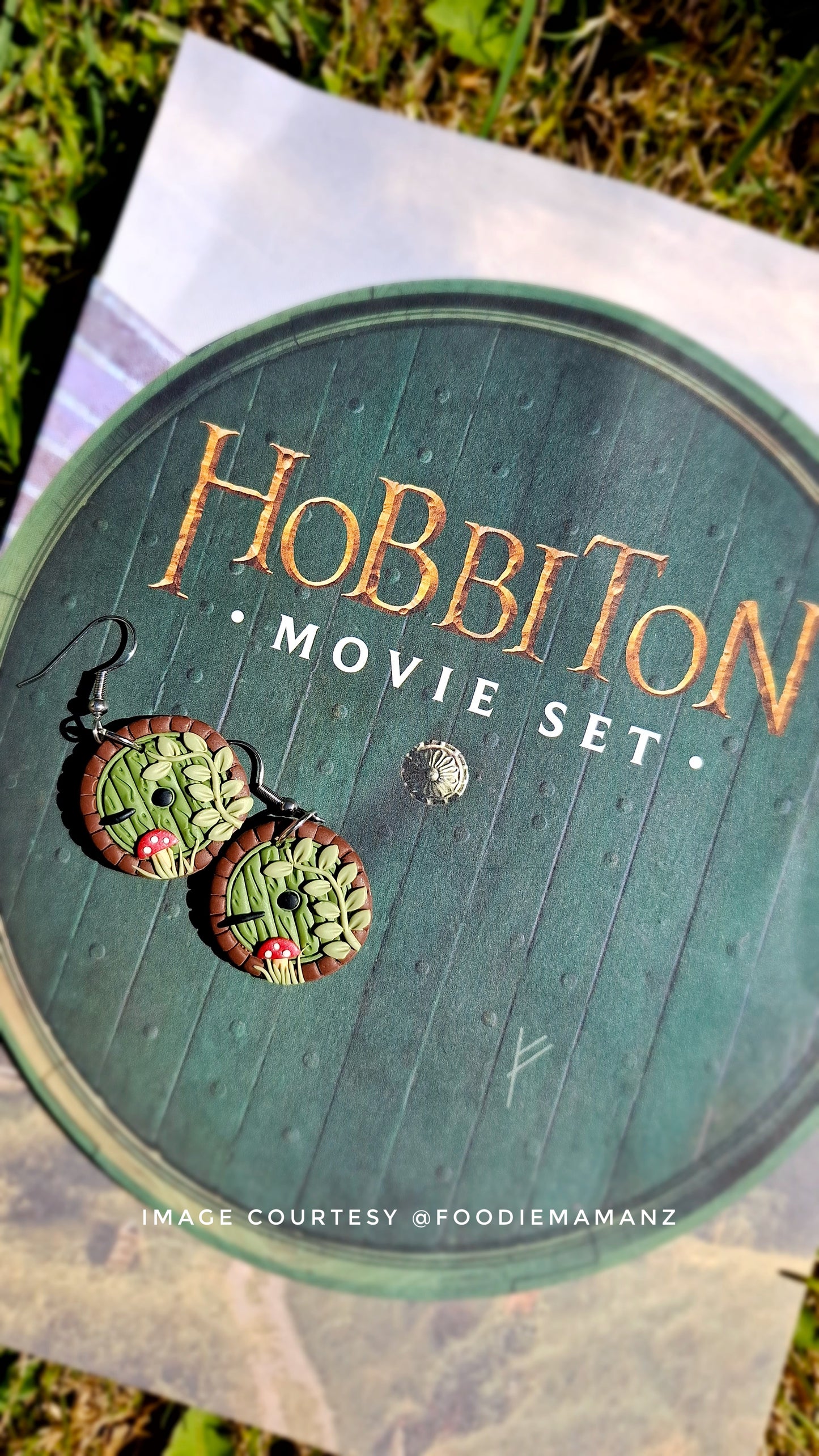 Close-up of a round green leaflet reading Hobbiton Movie Set, featuring silverstar.nzs Hobbit Door Polymer Clay Earrings. The unique handmade design showcases textured green hobbit doors with tiny red details, set against a grass-peeking backdrop.