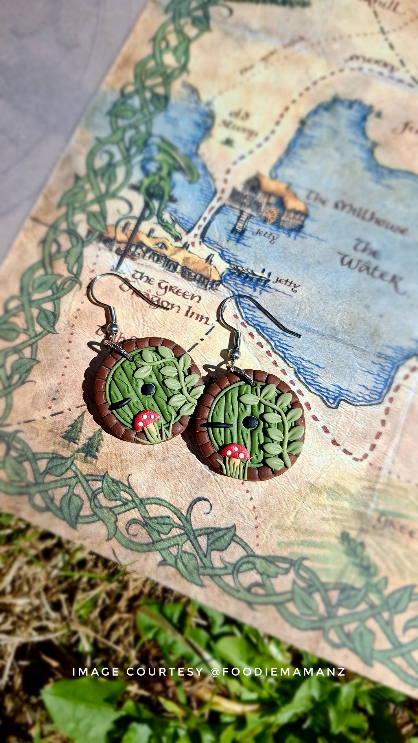 The Hobbit Door Polymer Clay Earrings by silverstar.nz feature intricate green and brown designs with round doors and detailed foliage patterns. These handmade, fantasy-inspired earrings rest on a map background. Image courtesy of @foodiemamanz.