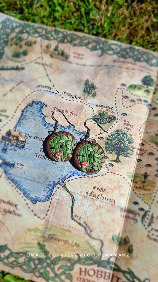 Two Hobbit Door Polymer Clay Earrings from silverstar.nz rest on a detailed fantasy map. Sunlight casts shadows, highlighting the maps texture and the earrings unique design. Grass appears at the top edge.