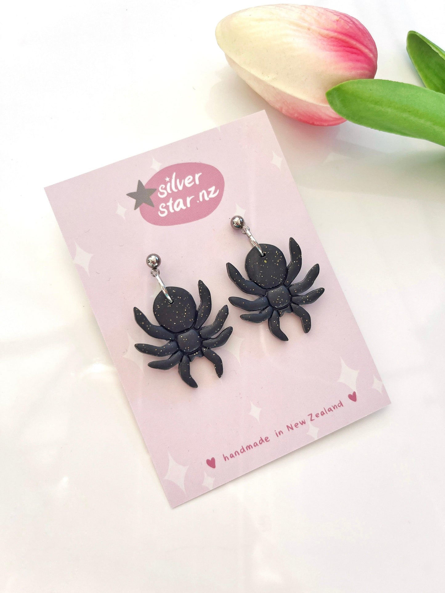 A trendy pair of black spider-shaped earrings, crafted from polymer clay with a glittery finish, is displayed on a pink card labeled as silverstar.nz. Accompanied by a pink and white tulip on the right, these stylish Spider Polymer Clay Earrings highlight their handmade origin in New Zealand.