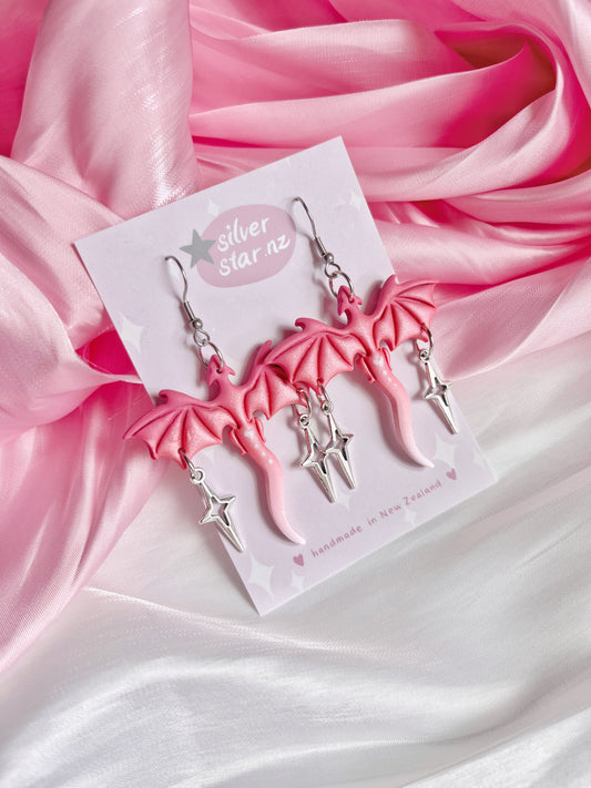 The Love Dragon Polymer Clay Earrings from silverstar.nz, featuring pink dragon wings with dangling silver stars, are set against a soft pink and white fabric backdrop.