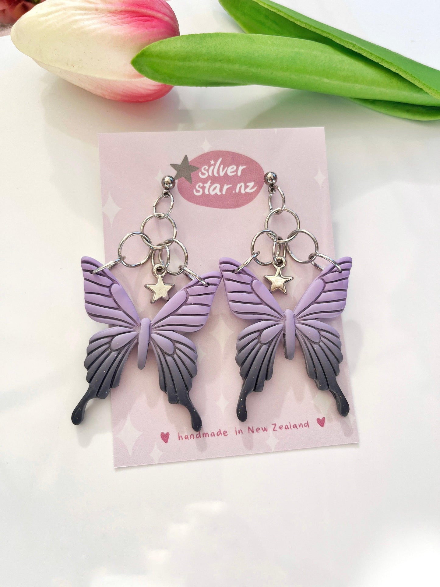 Displayed on a silver star.nz card is a pair of Statement Butterfly Polymer Clay Earrings, featuring purple butterfly designs with star charms and stainless steel hooks. A pink and white tulip rests above them on a glossy surface, enhancing the unique, handmade-in-New Zealand design of these earrings.