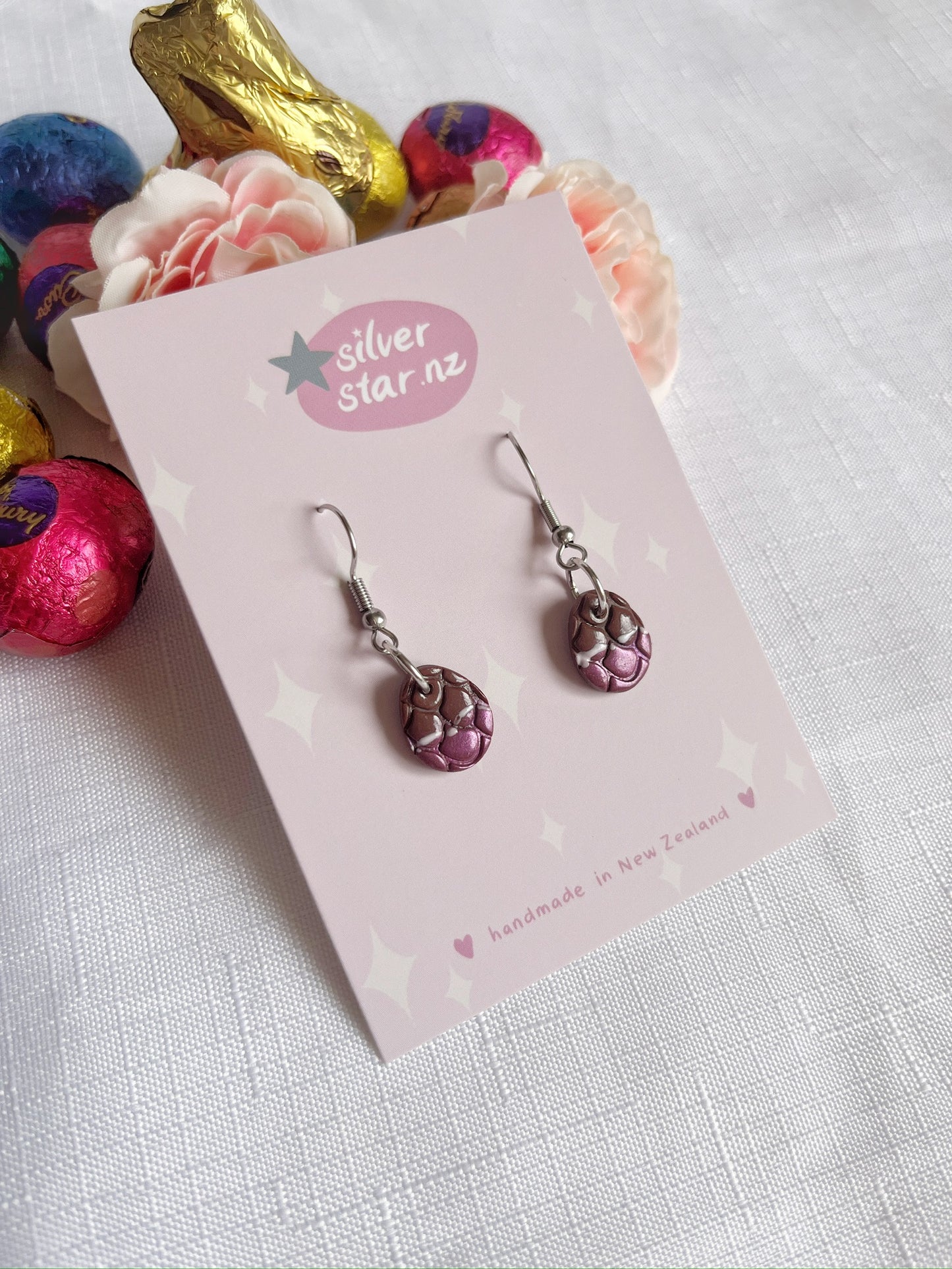 Opened Easter Egg Polymer Clay Earrings by silverstar.nz, featuring a unique handmade design reminiscent of purple dragon scales, are displayed on a card among vibrant chocolate eggs and flowers on a white surface.