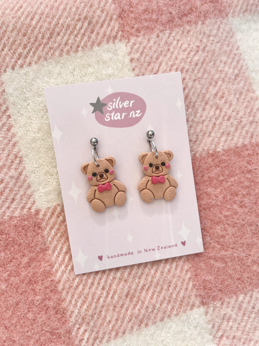 A pair of Teddy Polymer Clay Earrings, showcasing a delightful design of whimsical bears adorned with pink bows, is artfully attached to a card labeled silver star.nz. These earrings feature a distinctive funky style and are expertly crafted in New Zealand with stainless steel hooks. They are beautifully presented against a pink and white checkered fabric background.