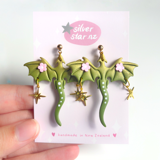A hand displays a card featuring Green Fae Dragon Polymer Clay Earrings, adorned with wings, pink flowers, and star charms. The card reads silverstar.nz and handmade in New Zealand, emphasizing their artisanal charm.