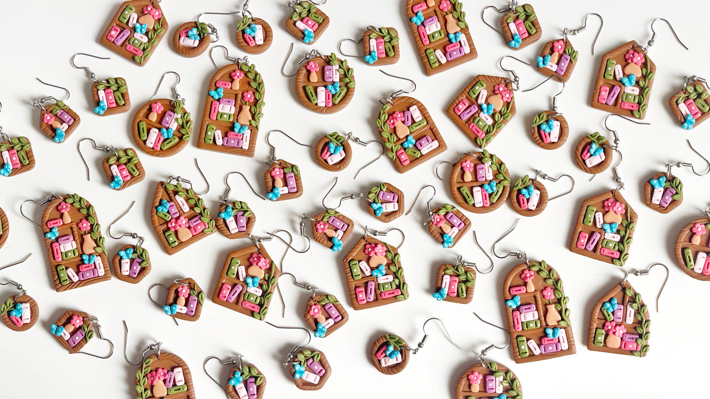 Introducing the Mini Hexagon Bookshelf Polymer Clay Earrings by silverstar.nz. These handmade, colorful earrings feature vibrant miniature houses with tiny flower accents. Each unique design varies in shape—round, oval, and house outline—and dangles from stainless steel hooks.