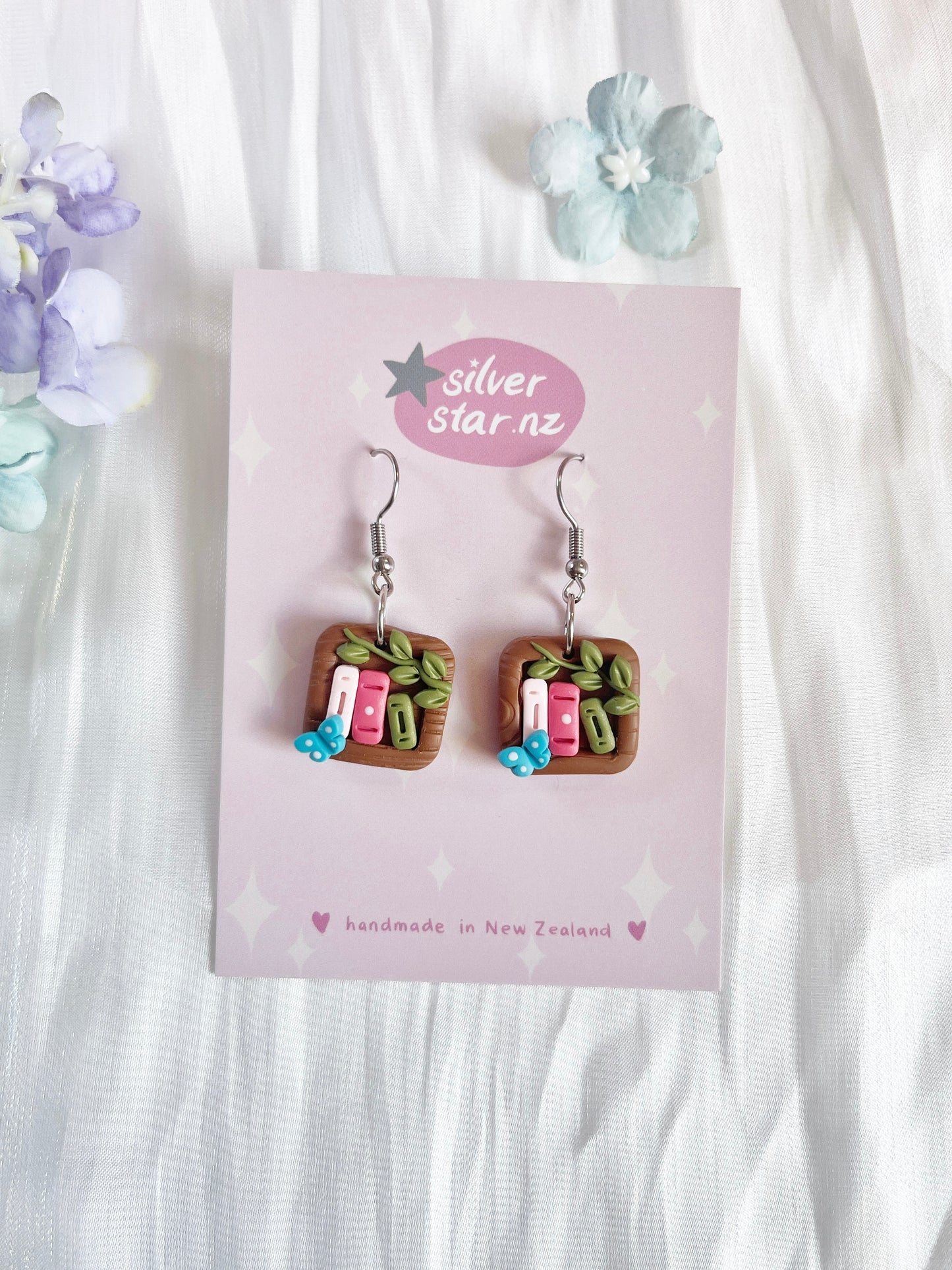 Explore silverstar.nzs Mini Square Bookshelf Polymer Clay Earrings, featuring funky miniature book designs with tiny leaves and blue flowers. With stainless steel hooks and presented on a floral pink card, these handcrafted earrings blend style and charm.