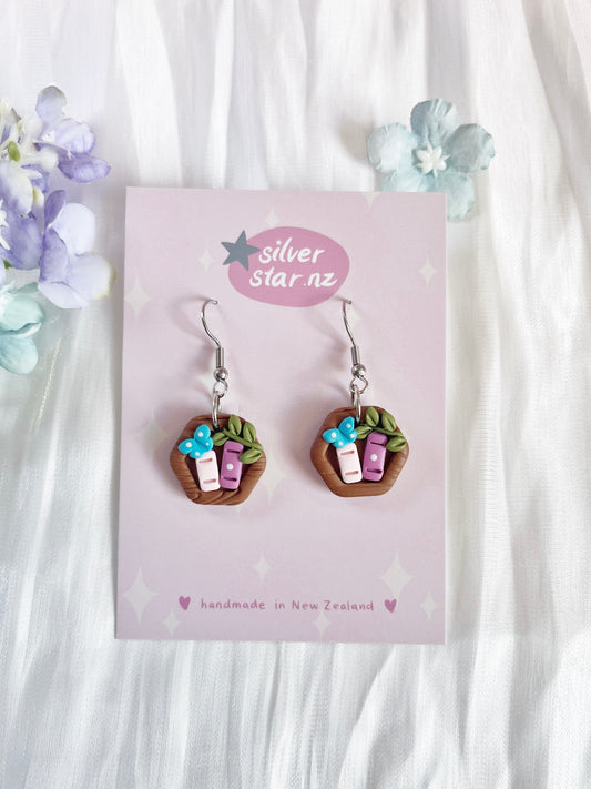 Mini Hexagon Bookshelf Polymer Clay Earrings feature baskets with handmade clay, pink and white objects, brown handles, and blue flowers. The card reads silverstar.nz with handmade in New Zealand. These unique earrings stand out against light fabric and pastel flowers.