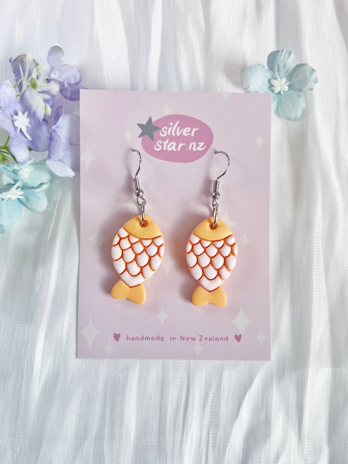 Fishy Polymer Clay Earrings, with orange and white scales, crafted uniquely from polymer clay by silverstar.nz, hang on a card on white fabric with pastel flowers. The card reads handmade in New Zealand.
