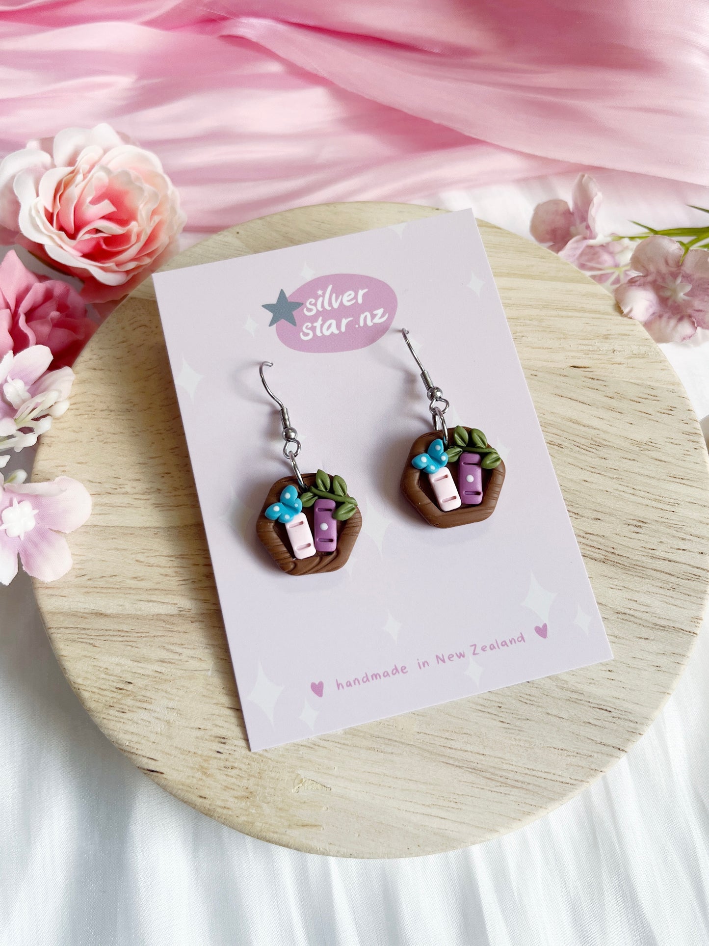 Mini Hexagon Bookshelf Polymer Clay Earrings feature tiny baskets with colorful books, a blue flower, and stainless steel hooks. Displayed on a silverstar.nz handmade in New Zealand card, theyre surrounded by soft pink and white flowers on a wooden surface.