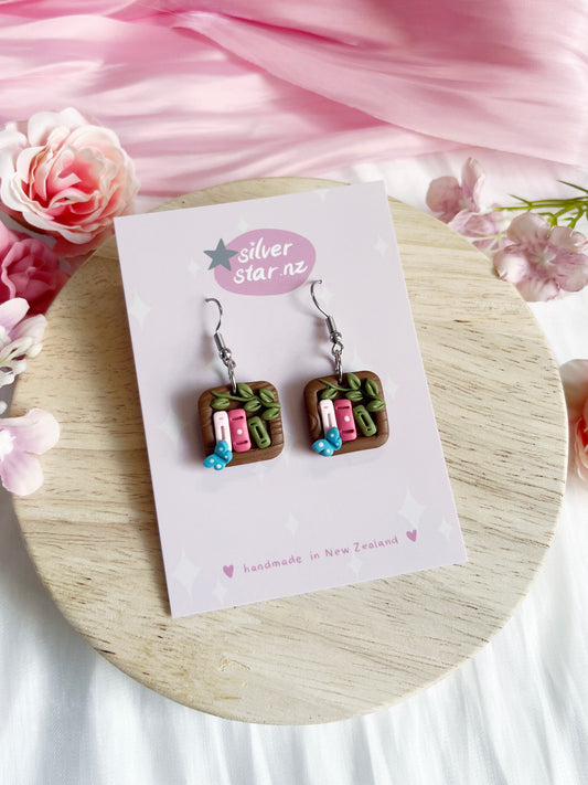 The Mini Square Bookshelf Polymer Clay Earrings by silverstar.nz feature a unique funky design with picnic tray motifs, tiny watermelon slices, and blueberries. They dangle from stainless steel hooks and are displayed on a card amidst pink flowers on a wooden surface.