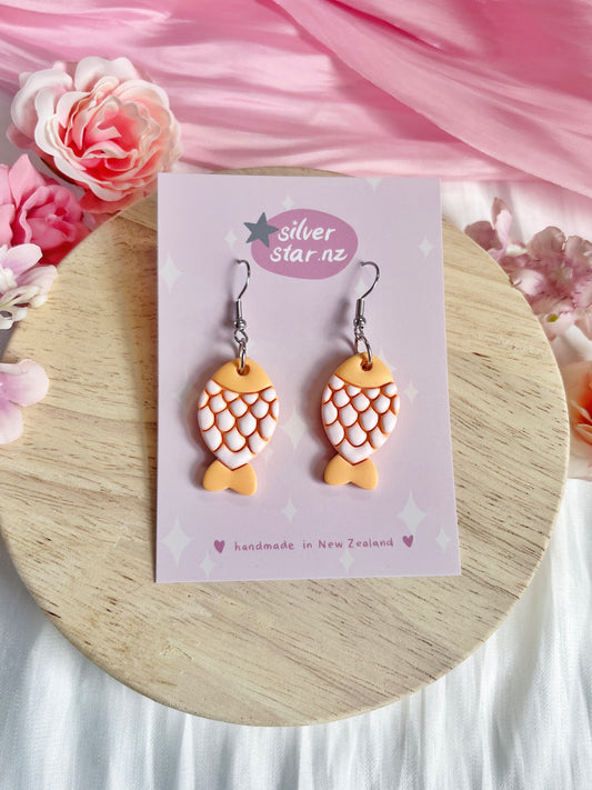 Fishy Polymer Clay Earrings by silverstar.nz, featuring a peach and white scale design, are displayed on a purple card stating silverstar.nz and handmade in New Zealand, set against a wooden surface with pink flowers.