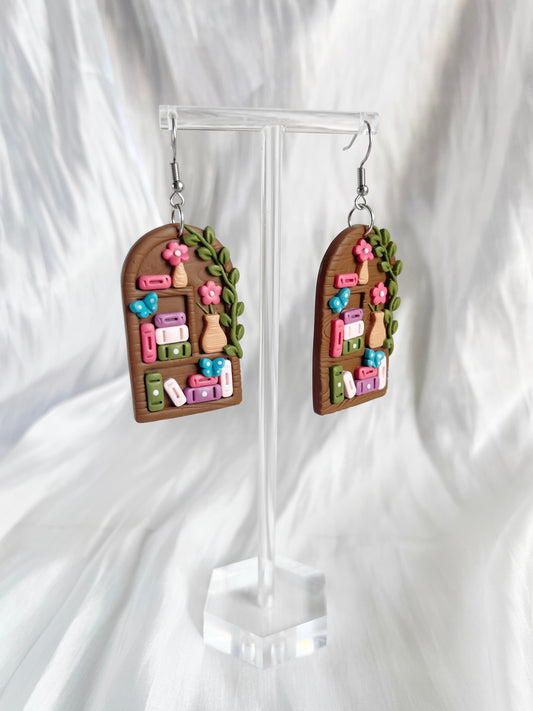 Tall Arch Bookshelf Polymer Clay Earrings by silverstar.nz feature arched bookshelf shapes with tiny colorful books, pink flowers, and green leaves on stainless steel hooks, displayed on a clear stand against soft white fabric.