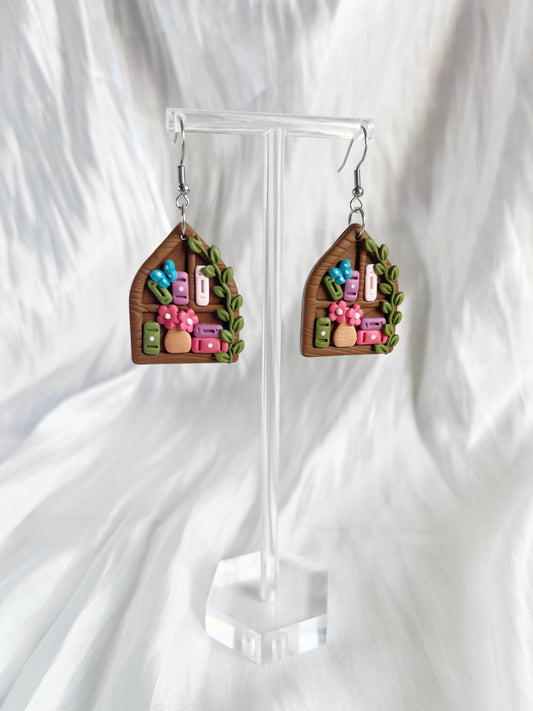 Pointed Arch Bookshelf Polymer Clay Earrings by silverstar.nz feature stainless steel hooks and showcase colorful books, pink flowers, and green leaves in a house shape. Displayed on a clear stand against soft white fabric.