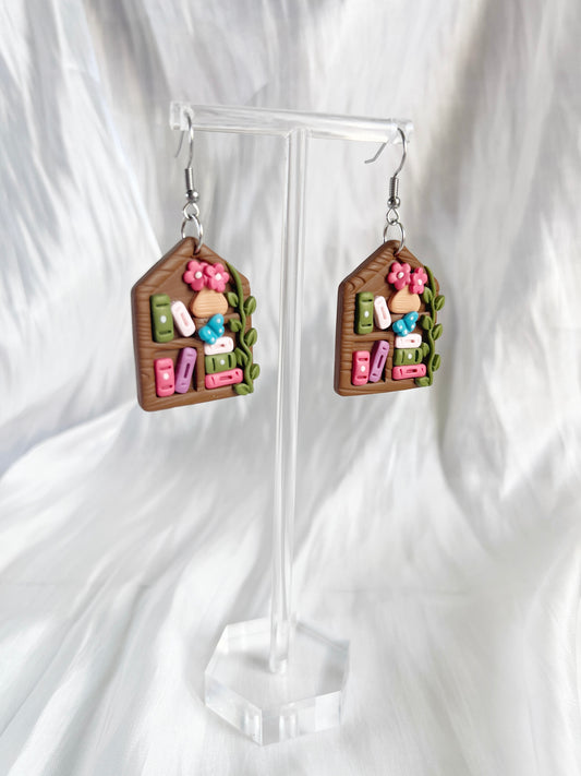 Cottage Bookshelf Polymer Clay Earrings by silverstar.nz feature miniature bookshelves with colorful books, pink flowers, and green leaves. Uniquely designed and displayed on a clear stand against a white fabric background.