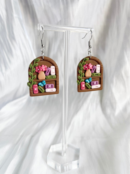 Round Arch Bookshelf Polymer Clay Earrings by silverstar.nz feature handmade bookshelf designs with colorful books, pink flowers, and green leaves. Displayed on a clear stand with stainless steel hooks for comfort against a soft white background.