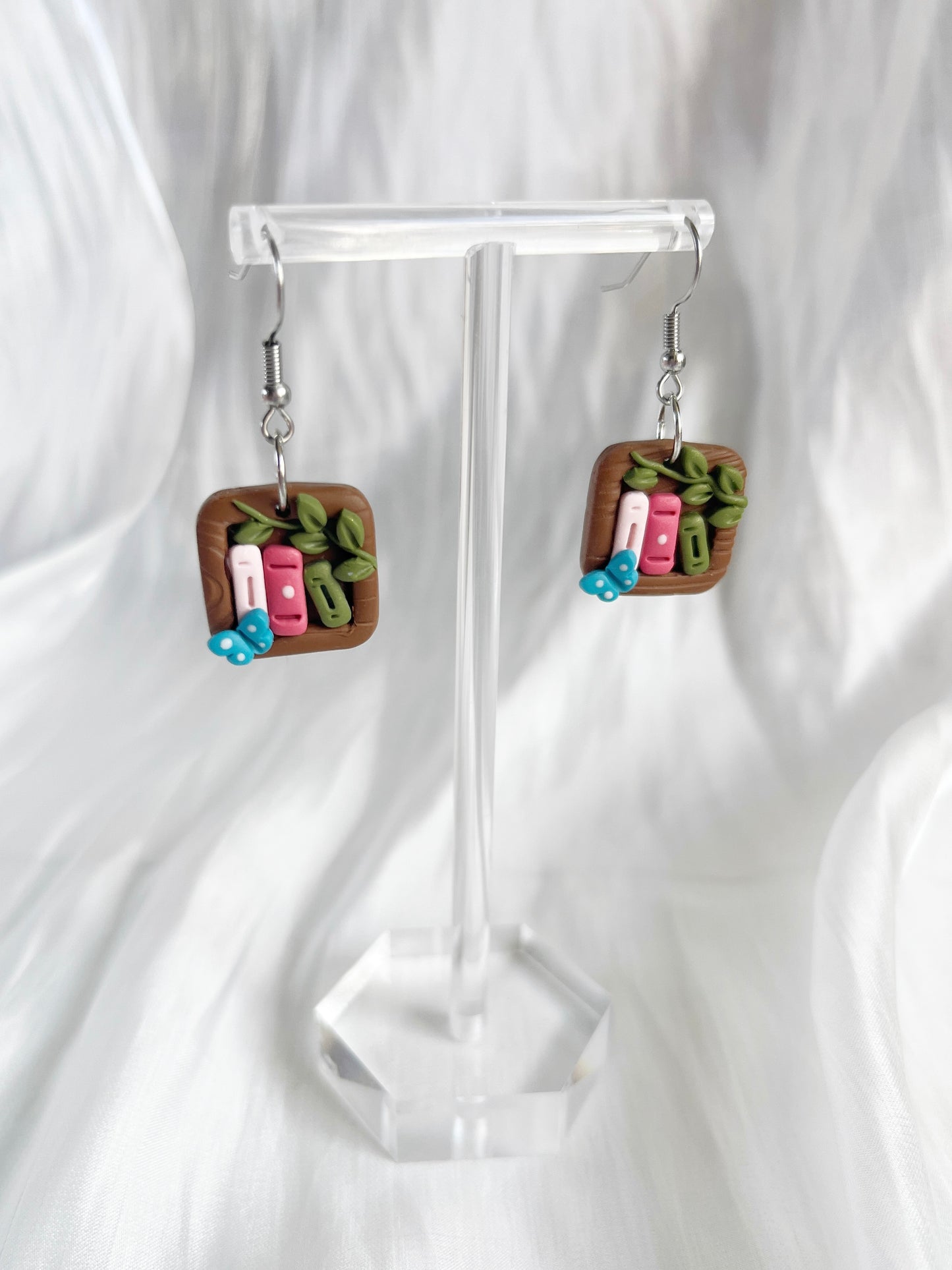 The Mini Square Bookshelf Polymer Clay Earrings by silverstar.nz feature square handcrafted designs with miniature book spines in pink, green, white, and a small blue butterfly on a clear stand against white draped fabric.