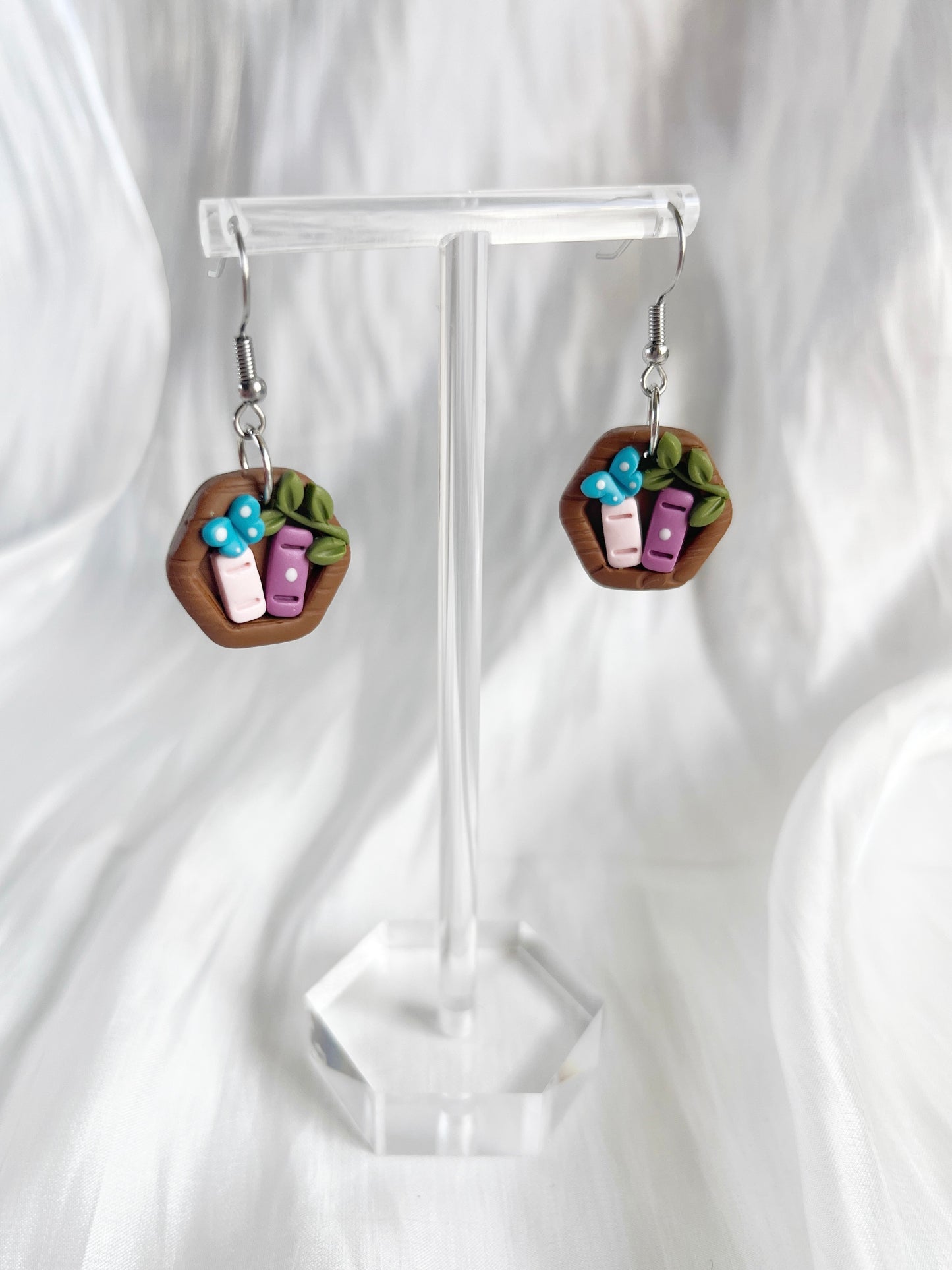 The Mini Hexagon Bookshelf Polymer Clay Earrings by silverstar.nz are showcased on a clear stand with a soft white background. Featuring tiny blue and purple book charms with green leaf accents, these whimsical earrings use stainless steel hooks for lasting wear.