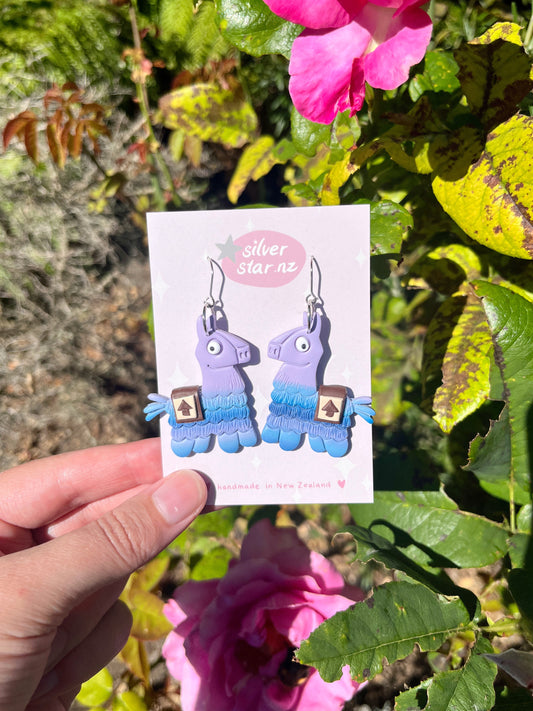 A hand holds a card with unique Fortnite Llama Polymer Clay Earrings featuring blue accents. The silverstar.nz card is adorned with pastel stars, set against a garden backdrop of pink flowers and lush greenery.