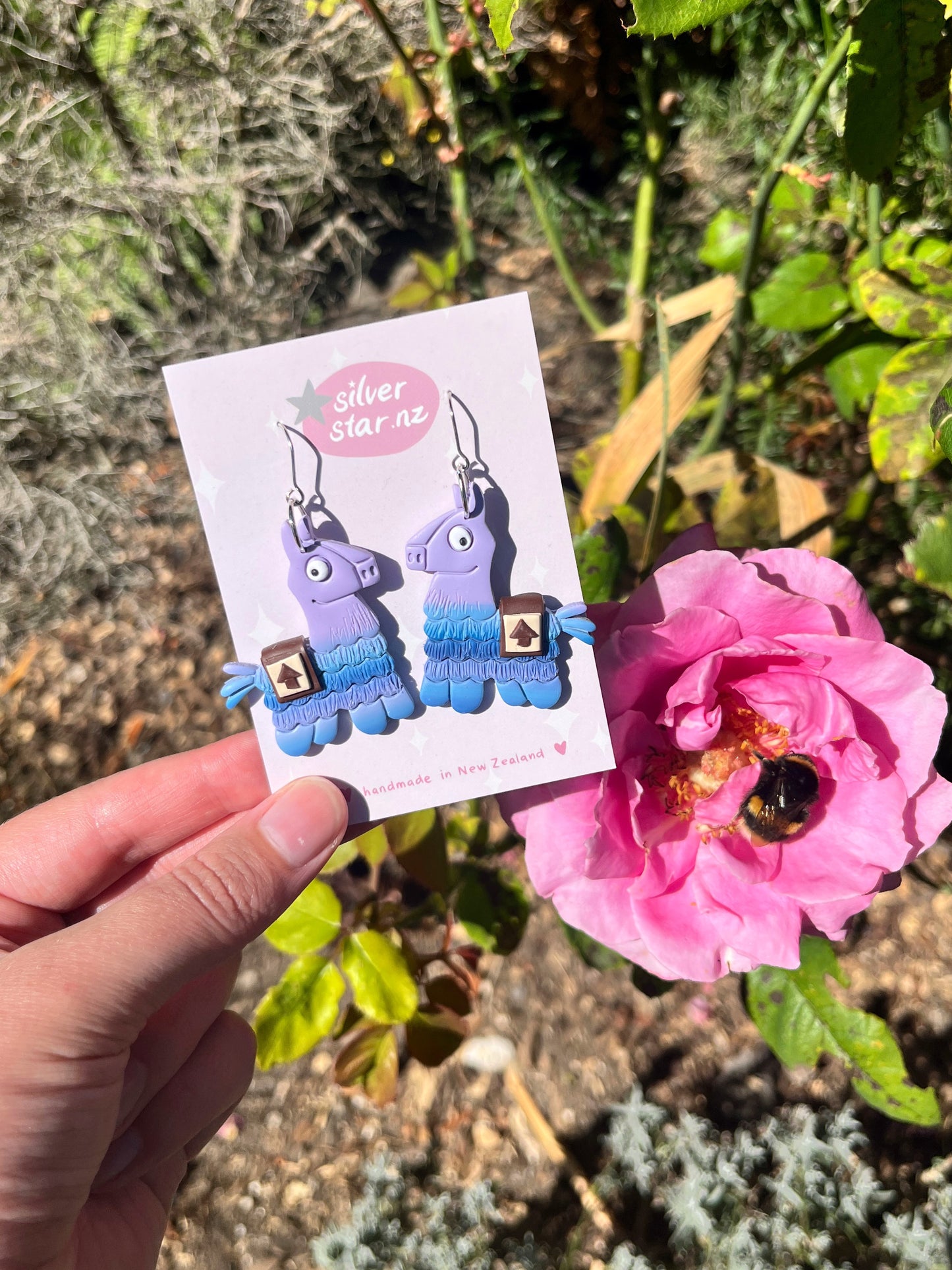 A hand showcases the Fortnite Llama Polymer Clay Earrings by silverstar.nz amid a garden. The earrings feature vibrant blue and purple hues with dangling fringes, while a nearby pink rose with a bee enhances the charm under the sunny sky.