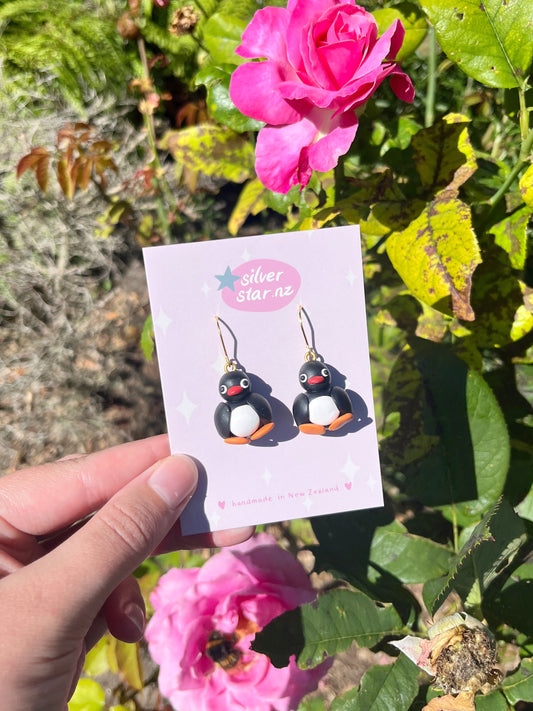 A hand holds a pair of Pingu Polymer Clay Earrings on a light purple card labeled silverstar.nz, set against vibrant pink roses and green foliage in bright sunlight.