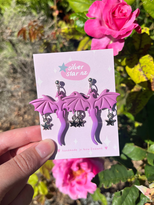 A hand holds a card displaying Purple Dragon Polymer Clay Earrings with star charms by silverstar.nz, photographed before bright pink roses and green foliage. The card says silverstar.nz and handmade in New Zealand.