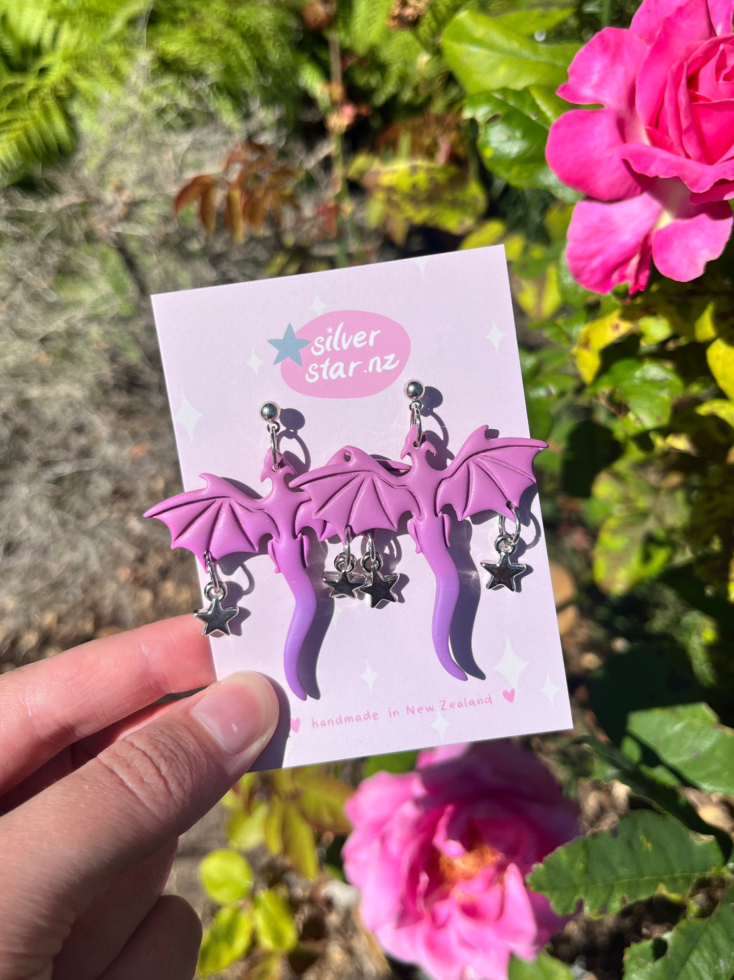A hand displays a card labeled silverstar.nz with three pairs of Purple Dragon Polymer Clay Earrings, handmade in New Zealand, featuring dangling silver stars, set against green leaves and a vibrant pink flower.