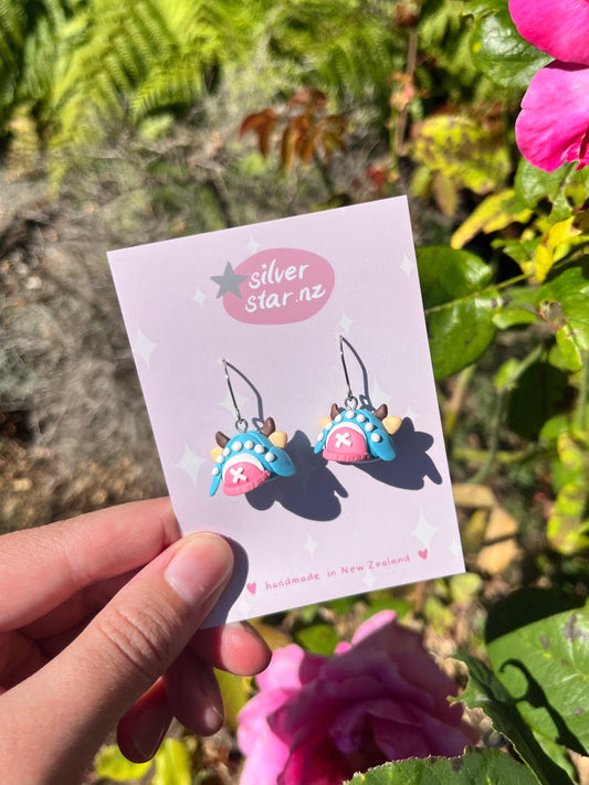 A hand holds a card with Chopper Hat One Piece Polymer Clay Earrings by silverstar.nz, displayed against green foliage and pink flowers in bright sunlight.