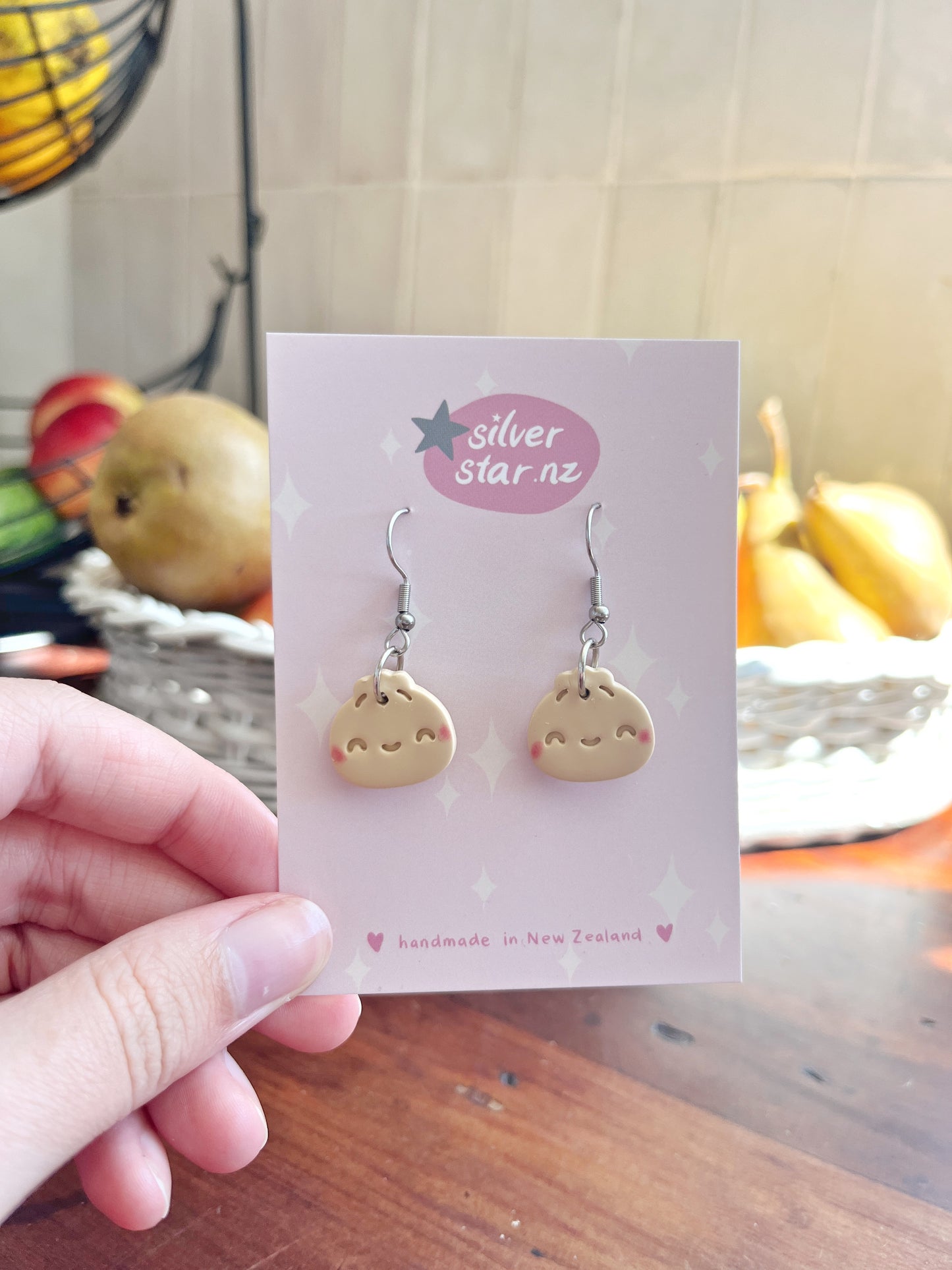 A hand displays a card with Bao Bun Polymer Clay Earrings, featuring cute dumpling faces and stainless steel hooks. The card reads silverstar.nz and handmade in New Zealand, while a fruit basket softly blurs in the background.