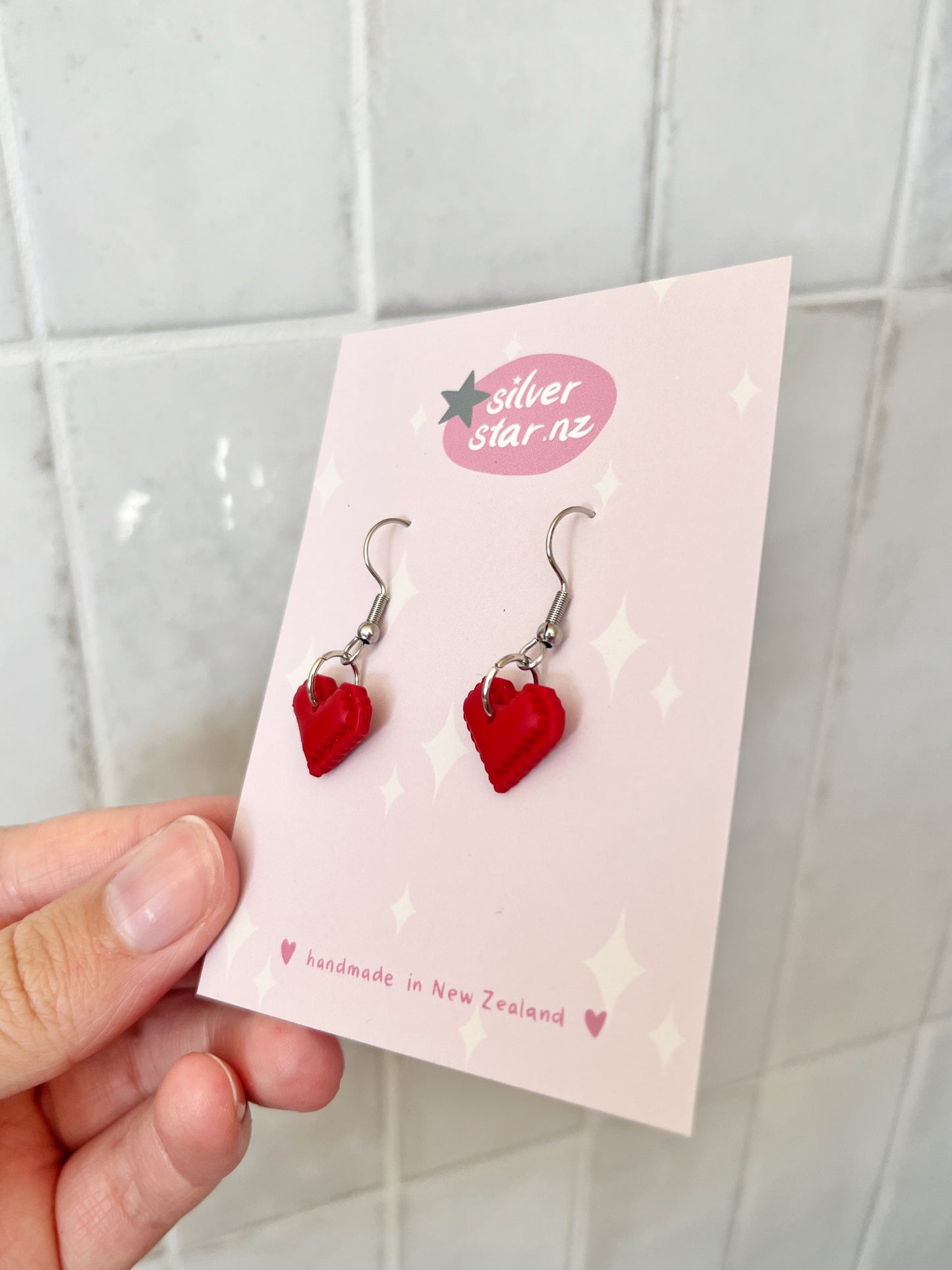 A hand holds a card with Pixel Heart Polymer Clay Earrings, featuring red hearts and stainless steel hooks. The card reads silverstar.nz and handmade in New Zealand, set against a pink background with star accents.