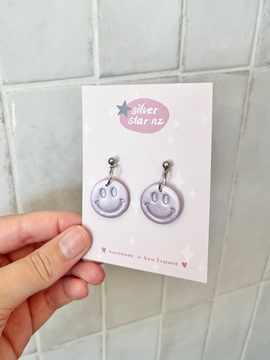 A hand displays a card from silverstar.nz featuring Smiley Polymer Clay Earrings with light purple smiley faces, labeled handmade in New Zealand against a light star-patterned background.