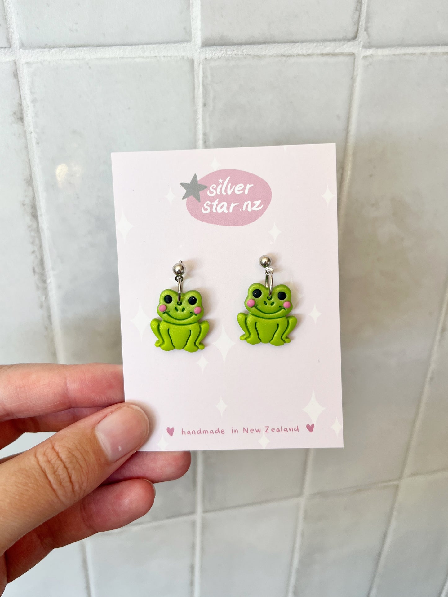 A hand holds a card reading silverstar.nz handmade in New Zealand, displaying funky Frog Polymer Clay Earrings with pink cheeks against a white tiled background.