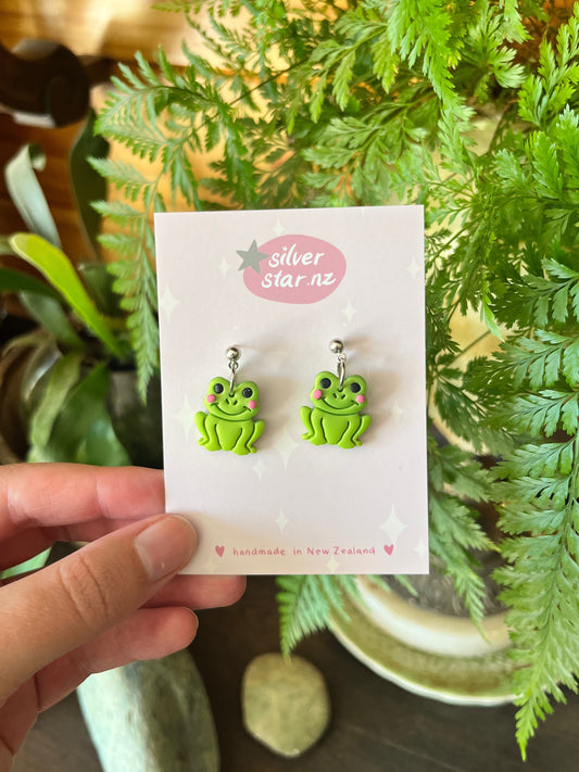 A hand displays Frog Polymer Clay Earrings from silverstar.nz, shown on a card with handmade in New Zealand in pink, held before leafy green plants.