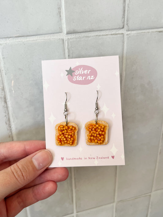 A hand holds a card from silverstar.nz displaying Beans on Toast Polymer Clay Earrings, handmade in New Zealand. The toast-shaped earrings with orange topping hang from stainless steel hooks, shown against a tiled background.