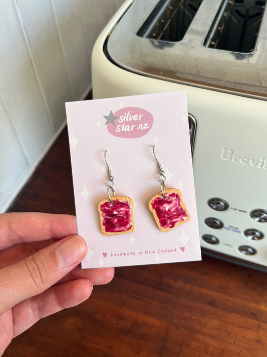 A hand holds Berry Jam on Toast Polymer Clay Earrings by silverstar.nz, displayed on a branded card before a white Breville toaster. These handmade earrings feature toast with red jam and dangle from stainless steel hooks.