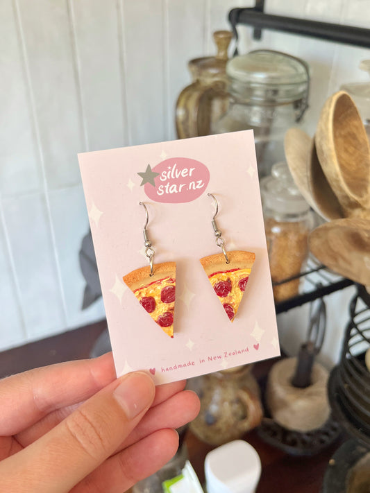 A hand displays Pizza Polymer Clay Earrings from silverstar.nz, crafted with stainless steel hooks. The earrings are shaped like realistic pizza slices and labeled handmade in New Zealand. Kitchen items are blurred in the background.
