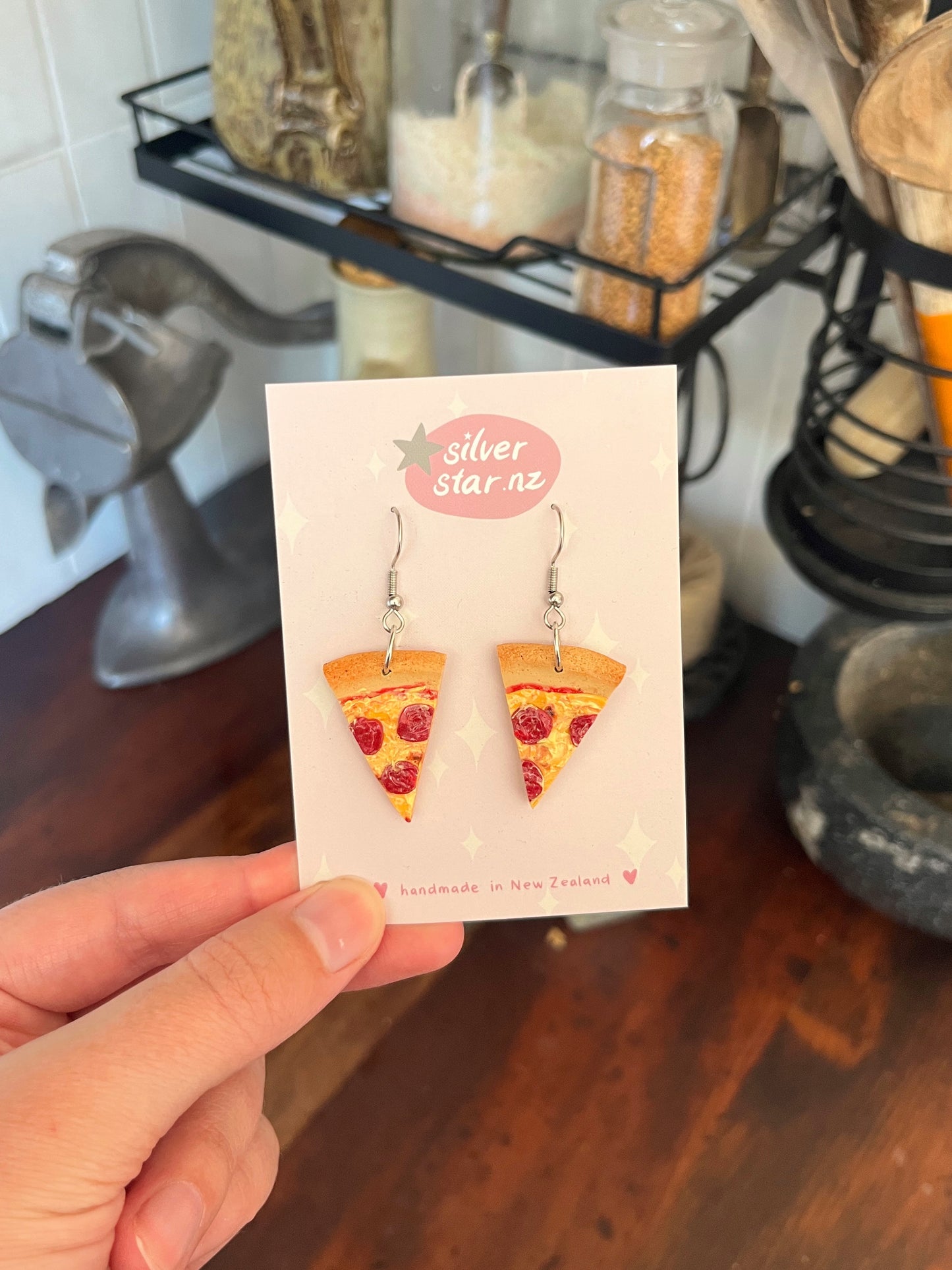 A hand holds a card from silverstar.nz displaying Pizza Polymer Clay Earrings, handmade in New Zealand with stainless steel hooks. Kitchen items are blurred in the background.