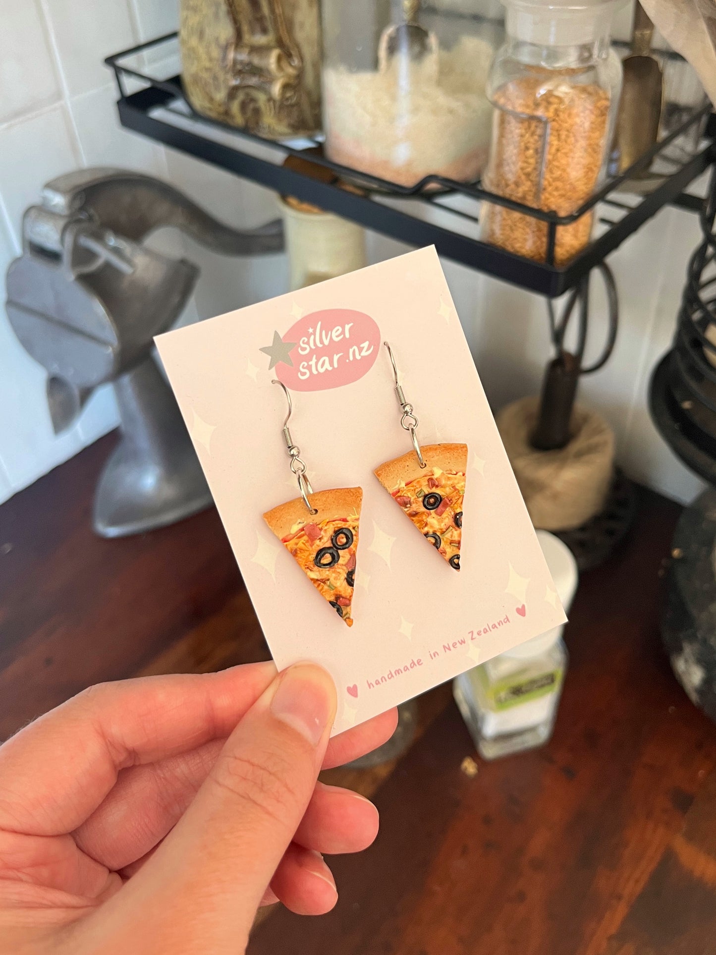A hand holds a card reading silverstar.nz handmade in New Zealand that displays Pizza Polymer Clay Earrings shaped like pizza slices with visible toppings, set against a kitchen background.