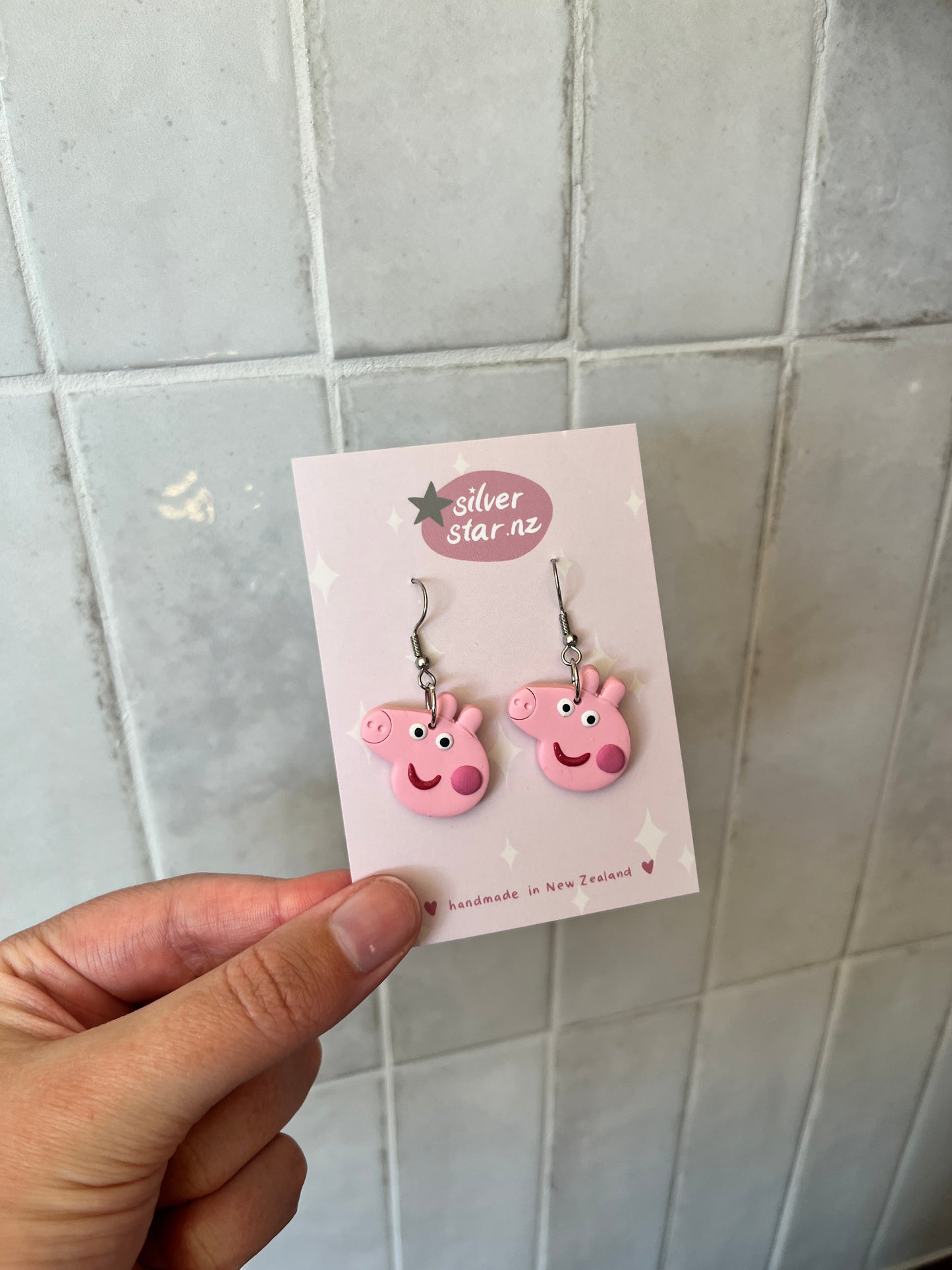 A hand holds a card displaying Peppa Pig Polymer Clay Earrings with smiling cartoon pig faces and stainless steel hooks from silverstar.nz, set against a background of pale, neutral-colored tiles.