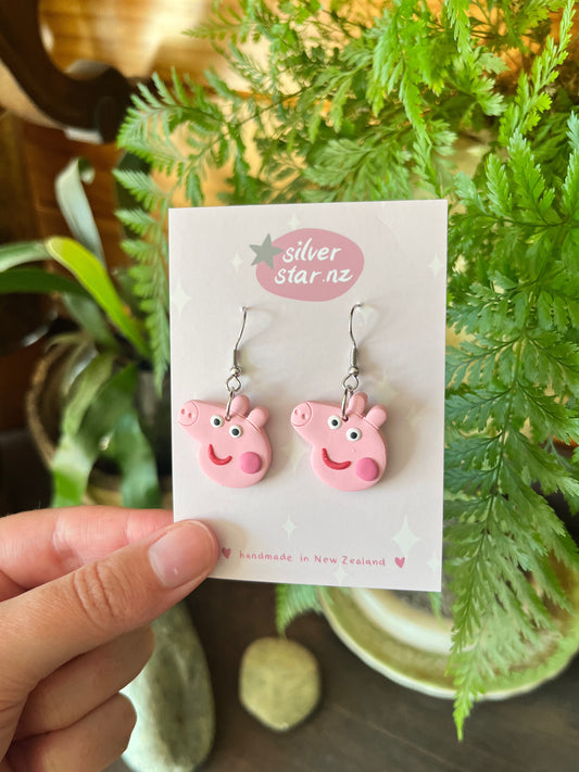A hand displays a card reading silverstar.nz with Peppa Pig Polymer Clay Earrings, handmade in New Zealand, shown against green plants and decorated with small star illustrations.