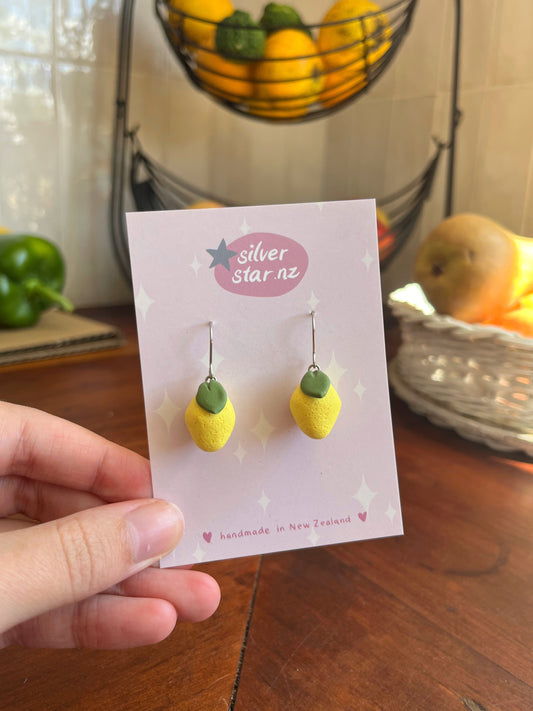 A hand holds a card featuring Mini Lemon Polymer Clay Earrings by silverstar.nz. Behind it, a basket overflows with colorful fruits and a green bell pepper on the kitchen surface. The card highlights their handmade in New Zealand craftsmanship and unique design.