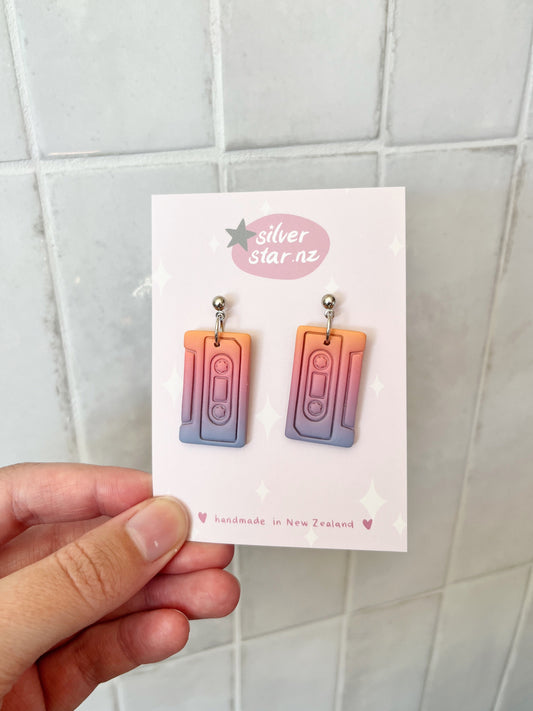 A hand displays Casette Polymer Clay Earrings by silverstar.nz—pastel ombre cassette tape shapes with stainless steel hooks, attached to a handmade in New Zealand branded card, shown against a tiled background.