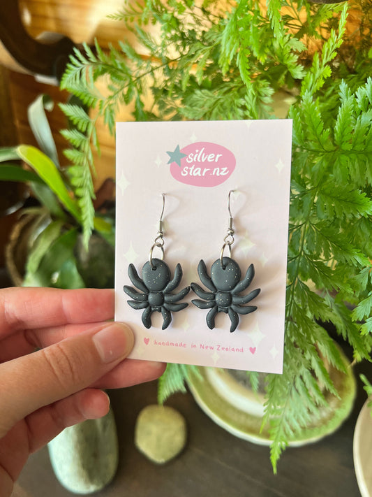 A hand holds a card with Spider Polymer Clay Earrings by silverstar.nz, labeled “handmade in New Zealand.” Lush green plants and smooth stones form the background.