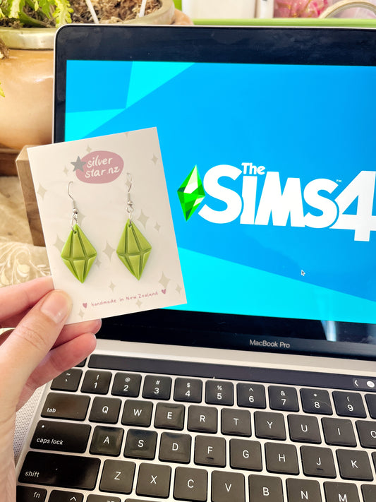 A hand holds Sims Plumbob Polymer Clay Earrings from silverstar.nz in front of a MacBook Pro with The Sims 4 logo. The handmade earrings, inspired by the game, are displayed on a branded card with lush green plants in the background.