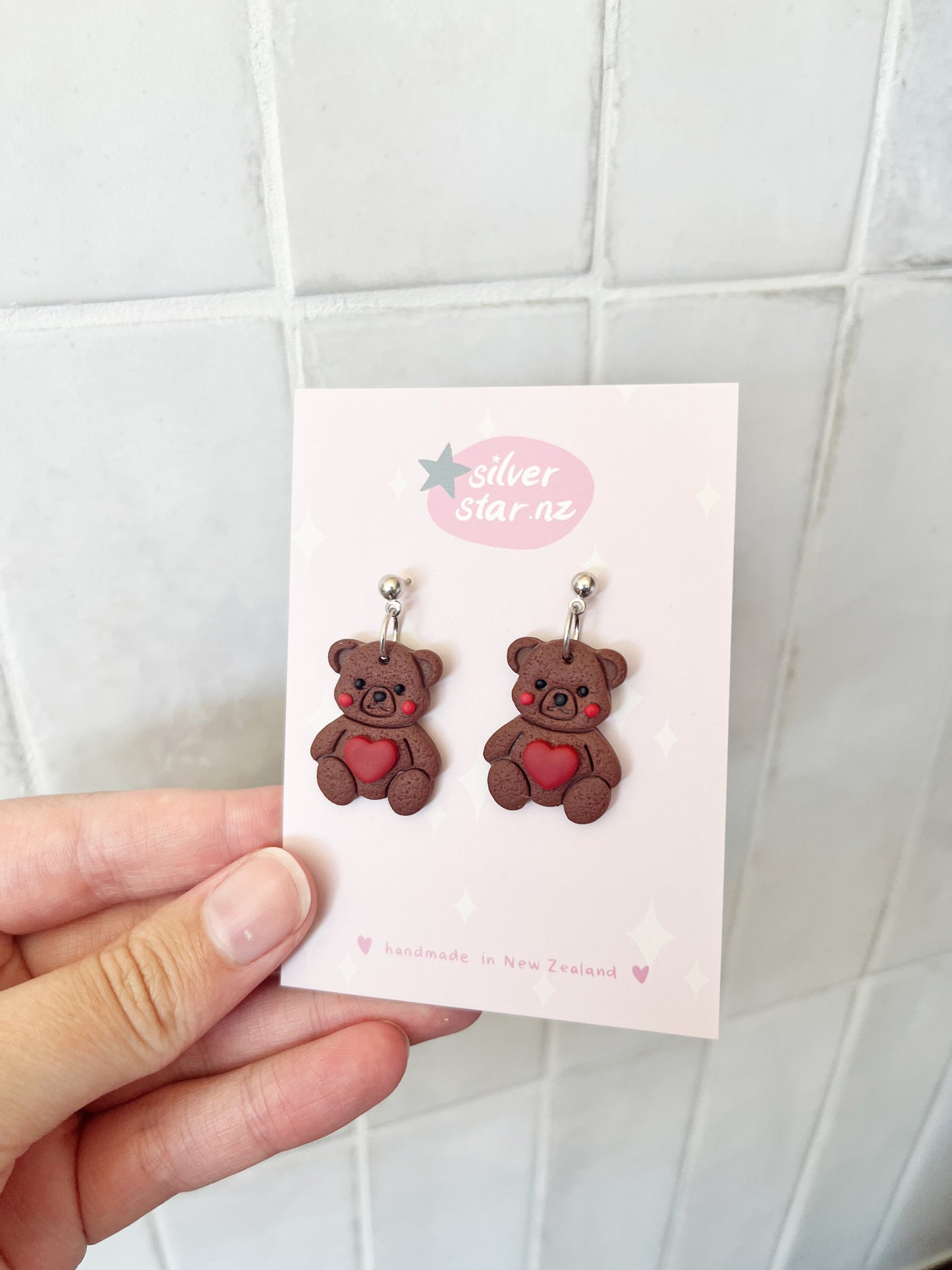 A hand holds a card with Love Teddy Bear Polymer Clay Earrings by silverstar.nz, each earring featuring a brown teddy bear holding a red heart. The card reads "handmade in New Zealand" and is shown against a tiled background.
