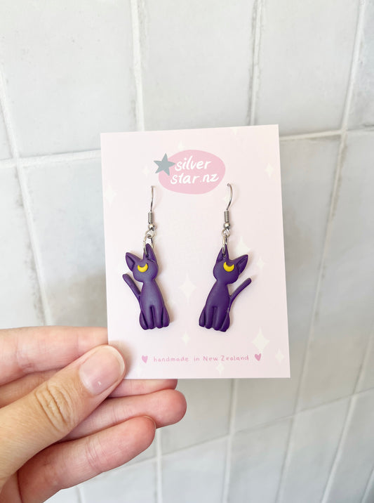 A hand holds a card displaying silverstar.nzs Luna Cat Polymer Clay Earrings, featuring funky purple cat designs with yellow crescent moons. Handmade in New Zealand, these earrings dangle from stainless steel hooks against a light-colored tiled background.
