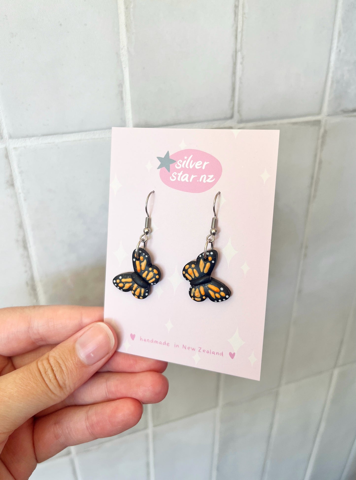 A hand holds a pink card with Monarch Butterfly Polymer Clay Earrings, featuring yellow and black butterfly designs. Handmade in New Zealand, they dangle from stainless steel hooks. The card displays silverstar.nz.