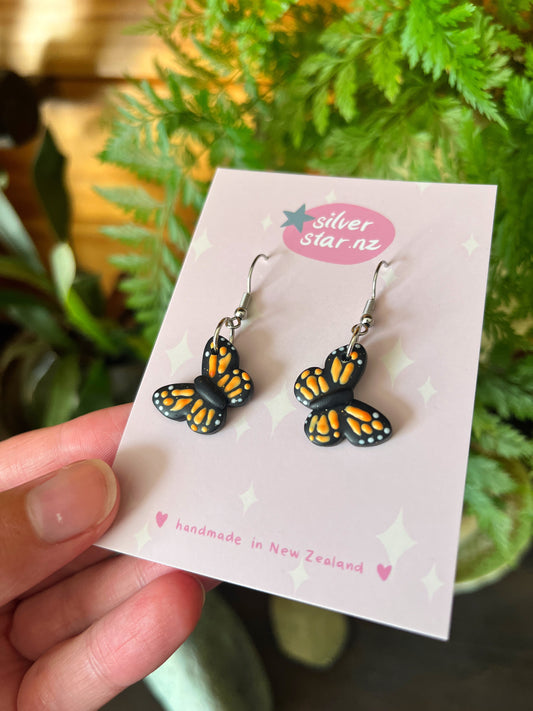 A hand holds the Monarch Butterfly Polymer Clay Earrings with orange, black, and white details on a silverstar.nz card, labeled handmade in New Zealand. These earrings have stainless steel hooks and stand out beautifully against lush green plants.