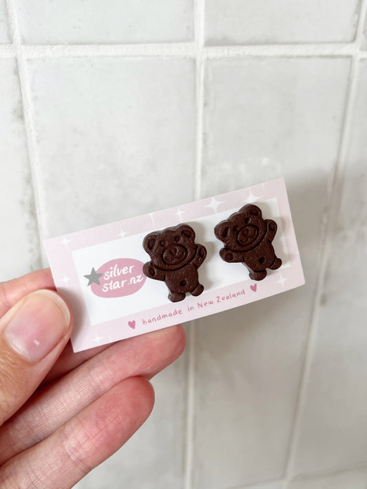 A hand holds a card featuring silverstar.nz Tiny Teddy Polymer Clay Stud Earrings, handmade in New Zealand. The card displays hearts and stars, set against a tiled background.